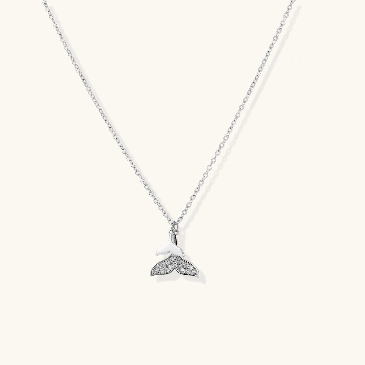 Delicate white gold plated whale tail pendant with pavé crystals on silver chain against cream background.