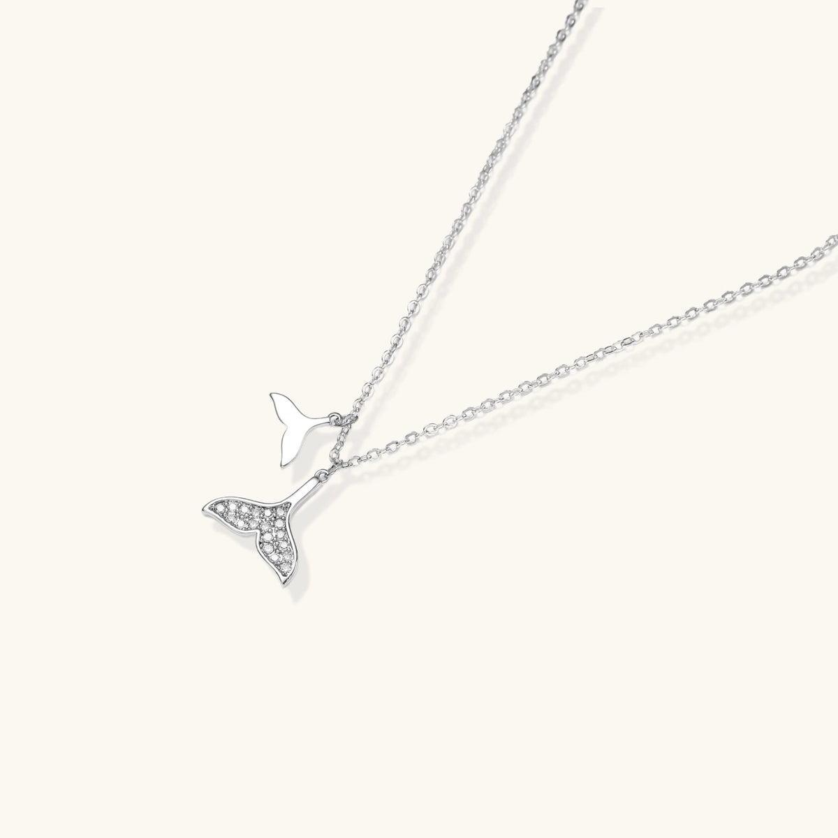 Delicate white gold plated whale tail pendant with pavé crystals on silver chain against cream background.